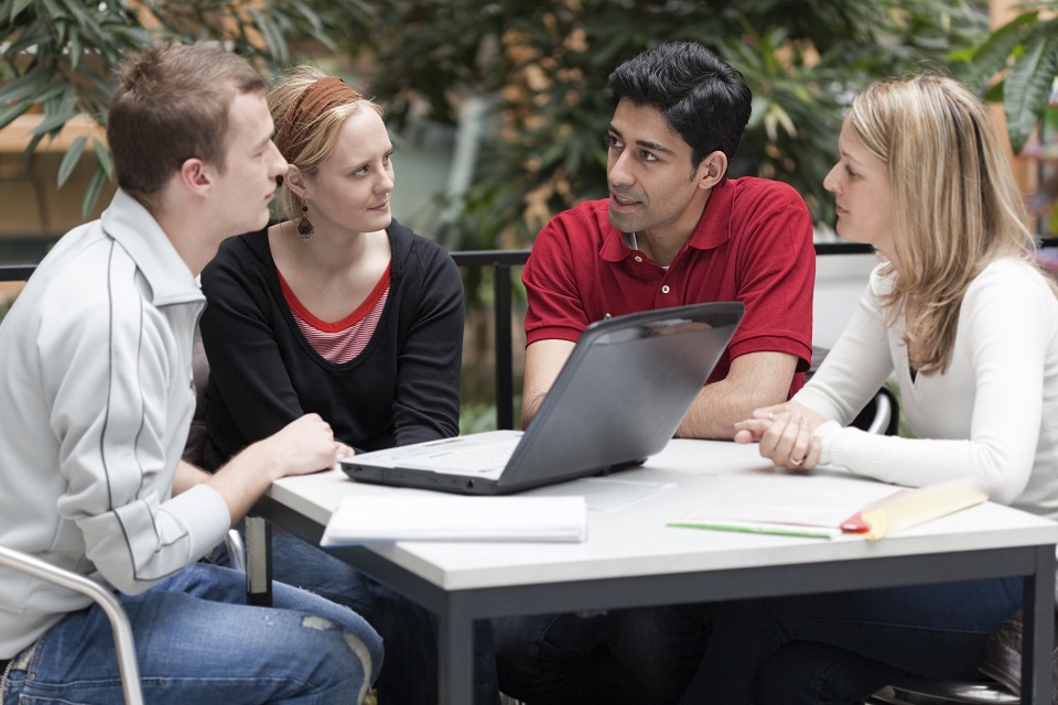 students sitting and talking around a laptop at a desk. 