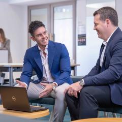 A man in a bright blue blazer is gesturing towards a laptop while looking at a second man in a dark blazer. 