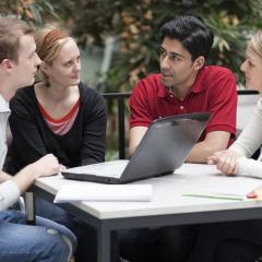 students sitting and talking around a laptop at a desk. 