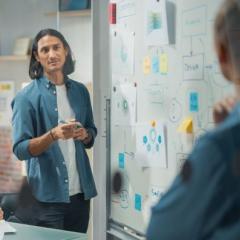 Man with long black hair stands at whiteboard looking at person in foreground 