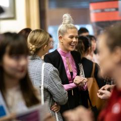 A young, blonde woman in a pink tshirt is smiling as she speaks with another person outside of frame