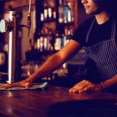 man wiping bar bench in foreground, bottles of wine on shelves in background. Image in a sepia tone.